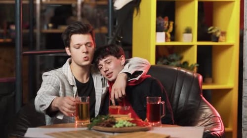 Dos jóvenes, muy borrachos, están bebiendo cerveza, sentados en una mesa en el bar y abrazándose