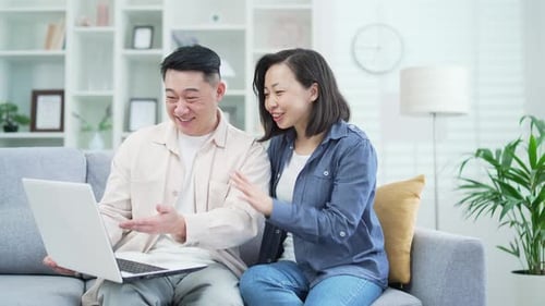 Happy asian couple shopping choosing products in online store using laptop, sitting on sofa