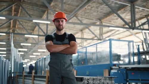 Worker in grey uniform and hard hat stands indoors in the factory with arms crossed