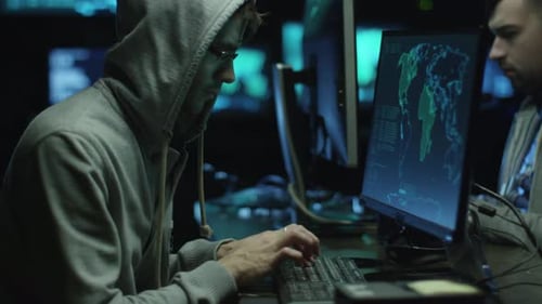 Dos piratas informáticos encapuchados trabajan en un ordenador con mapas y datos en pantallas de visualización en una oficina oscura