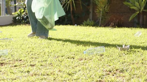 Mujer africana adulta que entra al patio con una bolsa verde y guantes doblándose, recogiendo botellas, ordenando el patio