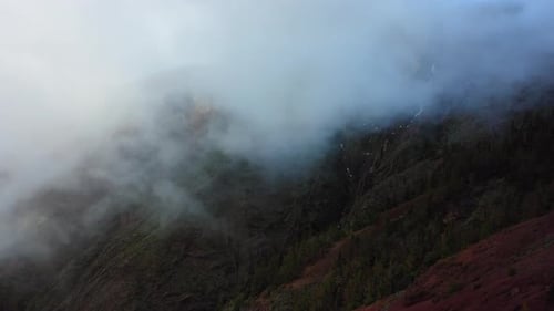 White air clouds descend down the mountainside into the valley. Aerial flying through mist in winter