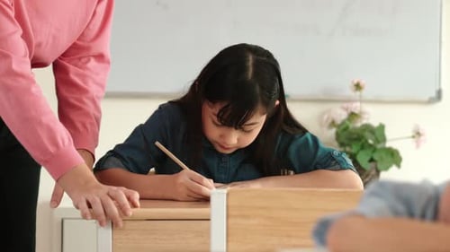 Girl writing at desk in classroom with teacher