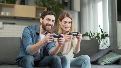 Happy adult man and woman playing with joysticks on play station exciting computer game while sit