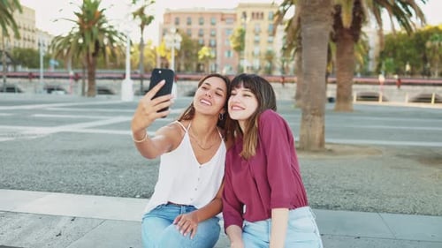 Two friends sitting on a bench laughing take a selfie with their smartphones to post on social media