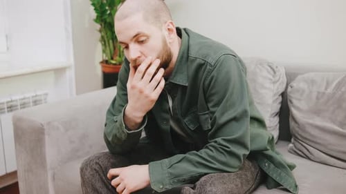 Sad Man Sitting on Couch in Contemplative Mood Indoors with Hand on Chin Reflecting on Thoughts in a