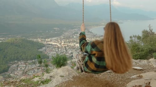 A panoramic view circles around the young woman swinging on swings over the cliff down.
