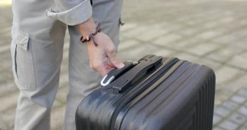 Man outdoors pressing suitcase release, extending handle, preparing to roll suitcase with bag strap