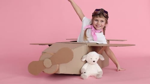 Glückliches kleines Mädchen im Pappflugzeug, Brille und Taschentuch spielt Pilot auf rosafarbenem Hintergrund