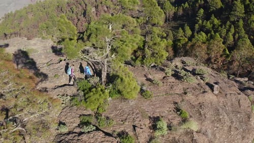 Pair of hikers walking an the top of a mountain surrounded by beautiful green forest. Aerial orbit s