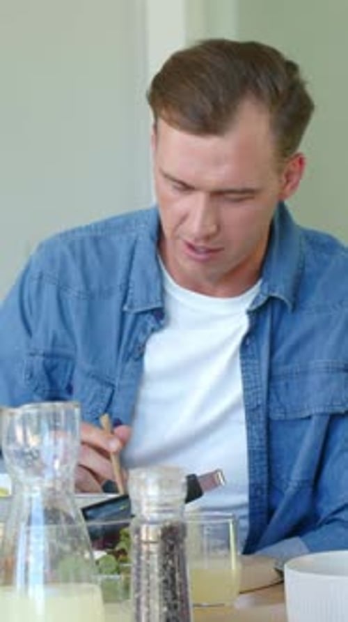 Vertical video: Eating adult man wearing blue shirt at table looking down at plate, reaching carafe