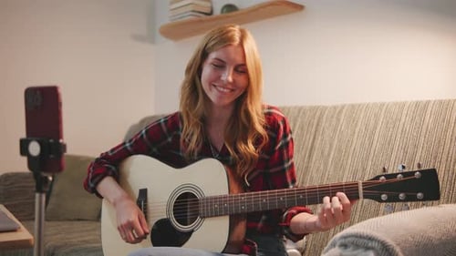 Woman playing guitar and talking to camera indoors