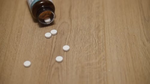 Jar of pills falls to the floor. The tables spill out across the wooden . Some of the white drugs ar