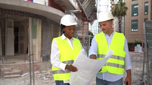 Two diverse engineers in helmets and vests reviewing a construction blueprint at a building site