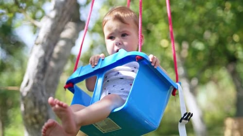 Sweet barefoot baby sits on a swing. Little kid having fun outdoors in the garden on warm sunny day.