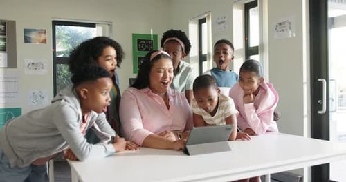 Female teacher with six children tapping tablet on white table in classroom sharing content