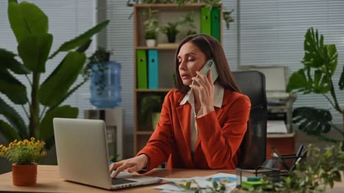 Older Adult Businesswoman Works at Green Office with Plants Woman Sitting at Desk Working on Laptop