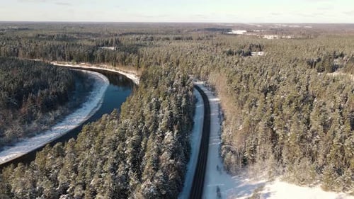 Aerial view of a winding river through a dense snow-covered forest in winter, with a curving road ru