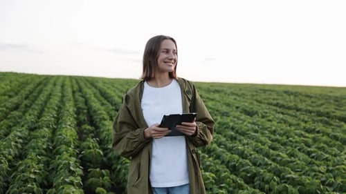 Smiling female agronomist walking at soybean plantation with digital tablet, examines plants and doe