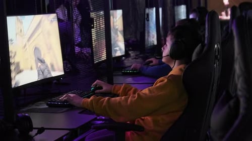 Teenager in Gaming Club Sits on Chair in Front of Monitor Playing Shooter Game