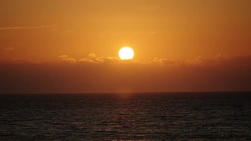 The sun sets above a blanket of clouds, casting intense golden hues over the calm Atlantic Ocean.