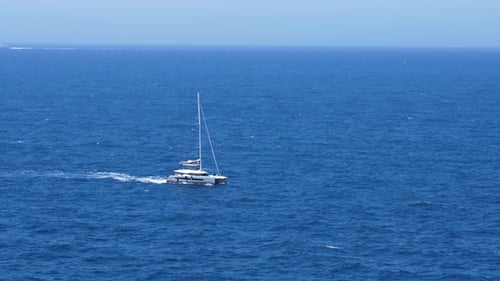 Un catamarán blanco solitario navega a través de un vasto océano azul con un cielo de niebla ligera en la parte trasera