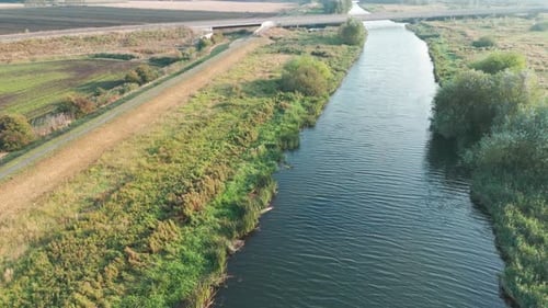 Aerial view of a serene river flowing under a bridge with adjacent green fields and a country road.
