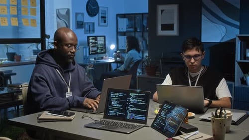 Two Multiethnic Young Male Data Analysts Working on Laptops in Dimly Lit Office