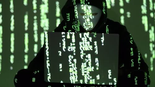 Masked Cybercriminal in Hoodie Facing Forward with Laptop and Green Matrix Code Reflections