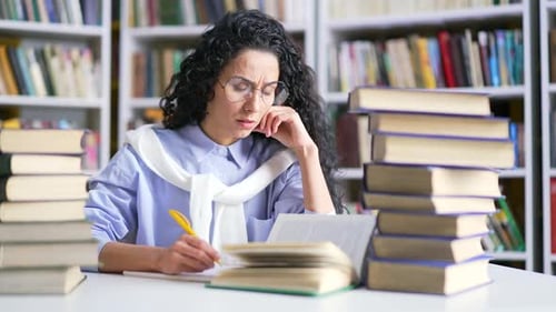 Female student studies by reading books, taking notes in campus library space. The girl learning
