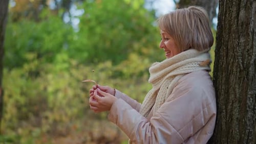 Frau lächelt, während sie das Herbstblatt in der Hand während eines friedlichen Waldmoments beobachtet