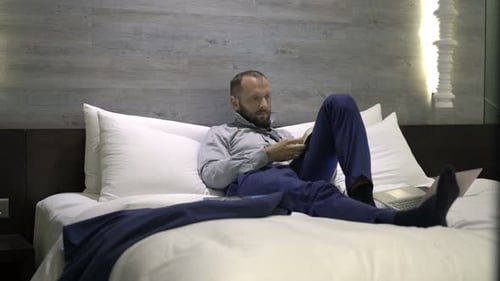 Businessman Reading Book Lying On Bed In Hotel Room
