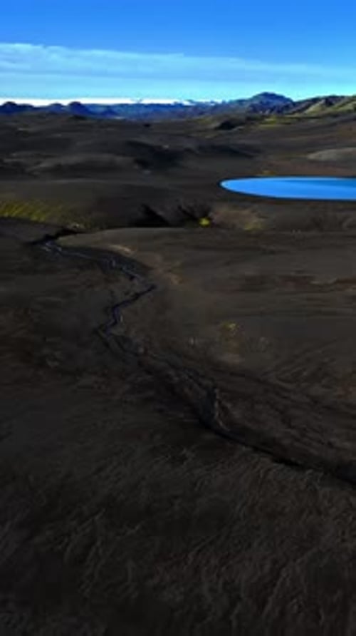 Vertical drone flyover of Iceland highlands: bright blue volcanic lake beside dark ash plains and a
