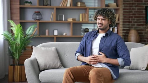 Upset young adult man feels stomach pain while sitting on sofa in living room at home. Sick
