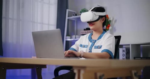 Asian woman using virtual reality headset while typing work on laptop in modern workspace.