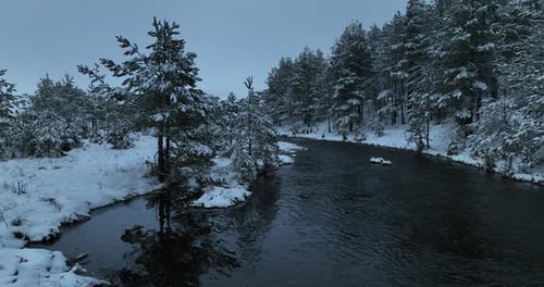 Eine Luftaufnahme eines gefrorenen Flusses, der durch schneebedeckte Wälder fließt, an einem bewölkten Sonnenuntergangshimmel
