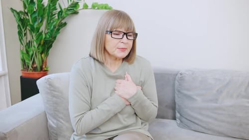 An Elderly Grayhaired Woman in a Cozy Sweater Sits on a Light Sofa with a Painful Expression on Her