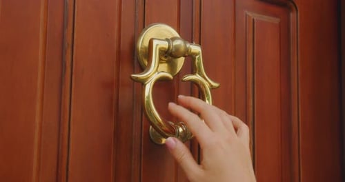 Hand holds ornate brass door knocker on a wooden door and knocks for the people living at home to op