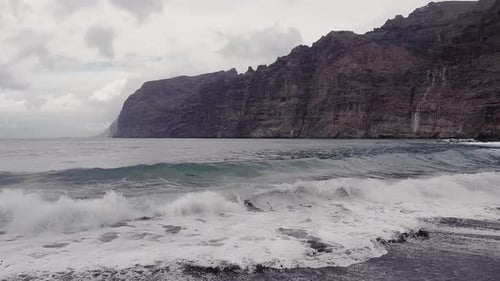 Moody cloudy ocean waves crashing on beach with tall rocky cliffs in distance. Aerial flying over se