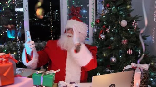 Santa Claus in a red suit, hat and beard chats fervently on phone, sitting next to pile of gifts