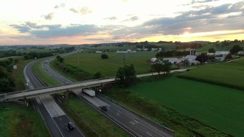 Cars and trucks, on highway under dramatic sunrise, sunset among green rural American farmland durin