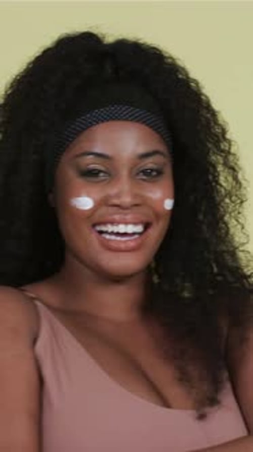 Vidéo verticale : Souriant à la caméra, une femme vêtue d'un bandeau lève les mains, étale de la crème sur le visage sur le plateau