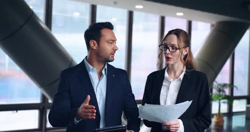 Two Businesspeople Discussing Paper Works While Walking In The Workplace Hallway. - medium shot