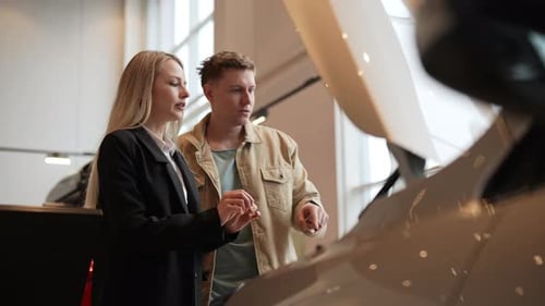 Female Car Dealer Describing Engine to Doubtful Male Buyer in Showroom Talking Gesturing Standing