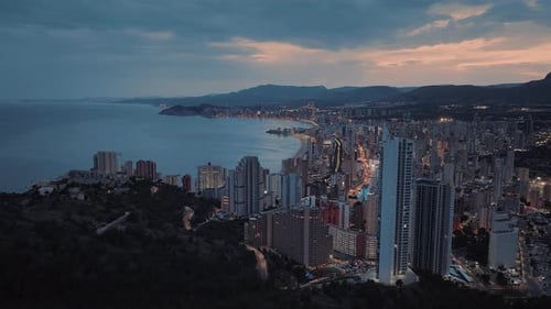 Modern seafront city with skyscrapers next to the rocky terrain. Calm blue ocean and dark clouds.