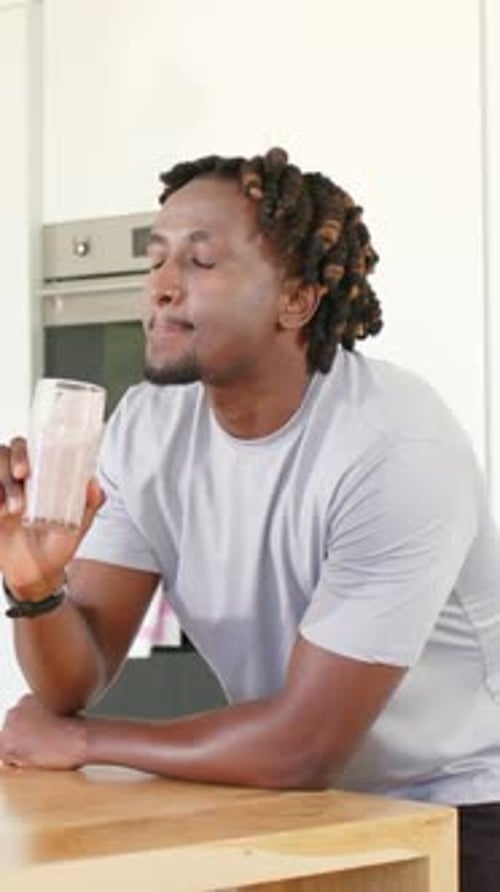 Vertical video: Lifting pink shake glass, man in gray tee sipping evaluating taste at wooden island