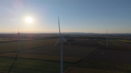 Several wind turbines rise above expansive farmland during sunset. The golden sunlight casts long