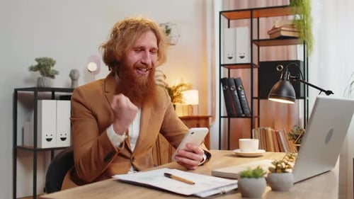 Bearded Man Excitedly Using Smartphone at Desk