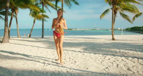 Smiling woman in bikini walks on white sand beach under palm trees by turquoise water