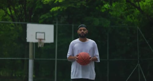 African American man standing on outdoor basketball court holding ball, looking straight ahead with